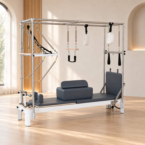 A Pilates reformer machine with a chrome steel frame, dark gray padded bed and headrest, and hanging white and black straps, photographed in a bright indoor studio with wood flooring and beige walls.